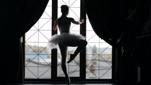 Ballerina in bwhite ballet tutu dress dancing classical pas in class near the window. Alone warming up before performance. Slow motion.