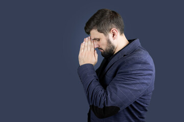 a stylish man with a beard in a blue suit stands sideways and holds his hands in front of his face