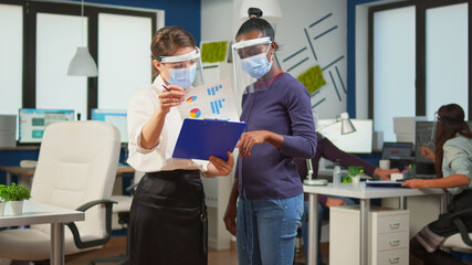 Company manager with visor against coronavirus explaining to african colleague project plan standing in front of desk holding clipboard. Multiethnic business team working respecting social distance.