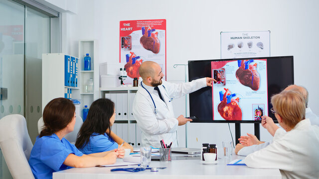 Experienced man doctor analysing heart issues image together with cvalified colleagues in meeting room, pointing on monitor. Doctors discussing diagnosis about treatment of patients
