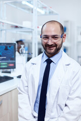 Naklejka na ściany i meble Young biochemistry scientist wearing protective glasses smiling at camera. Serious expert in genetics in lab with modern technology for medical investigations with african assistant in the background.