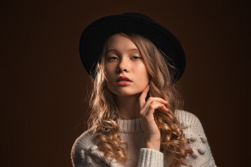 Charming blonde teen girl in black hat posing in studio on brown background