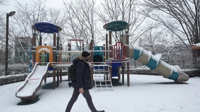 A snowy park in Seoul is quarantined off due to covid19. Man with jacket and backpack enters FG from left and exits to right. 4K 60fps tripod. Leafless trees in BG. Snow completely covers the ground.