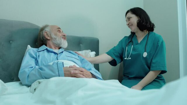 Cheerful nurse talking to senior patient, lying in bed, medical care, companion