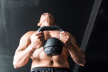Looking up at a well toned and fit shirtless man holds a heavy kettlebell ready to do an intense workout. Industrial gym setting.