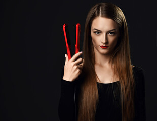 Obraz premium Serious woman young professional stylist with silky long hair wearing black clothes stands holding red hair straightener in hand at copy space over dark background. Haircare, beauty, wellness concept