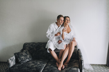 A beautiful bride with curly hair in a white robe and a long veil and a smiling groom are sitting on the sofa and drinking champagne from a glass glass. Wedding portrait in the morning at the hotel.