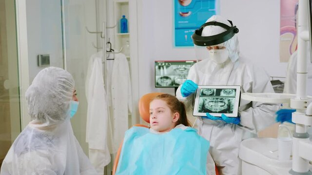 Dentist with face shield explaining panoramic mouth x-ray image to mother of kid patient during global pandemic. Stomatologist talking with woman wearing suit, coverall, protection suit, mask, gloves
