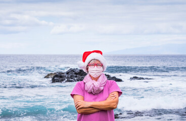 Obraz premium Senior woman in Santa hat wears a surgical mask due to coronavirus infection and enjoys the winter sea looking at camera. Horizon over water behind her