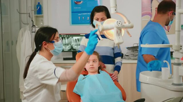 Woman orthodontist lighting the lamp examining little patient using sterile dental tools in stomatological clinic. Dentistry doctor speaking to kid sitting on stomatological chair, girl opening mouth
