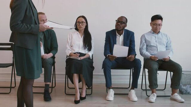 Full shot of four nervous diverse job applicants sitting on chairs in hallway, waiting for their turn, unrecognizable manager inviting young African man