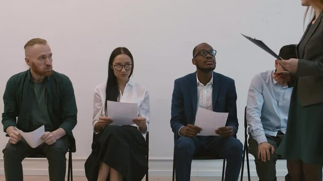 Medium long shot of four multiethnic impatient people sitting on chairs in hallway, holding resumes in hands, cropped HR manager inviting young Asian female candidate for interview, leaving men there