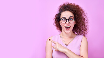 Portrait of amazed caucasian curly girl wearing glasses points finger to the side on copy space...