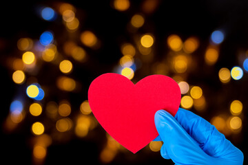 Fototapeta premium Valentine's day concept. A hand in medical blue gloves holds a red heart shape on a black background, glowing bokeh, copy space. Love, health concept