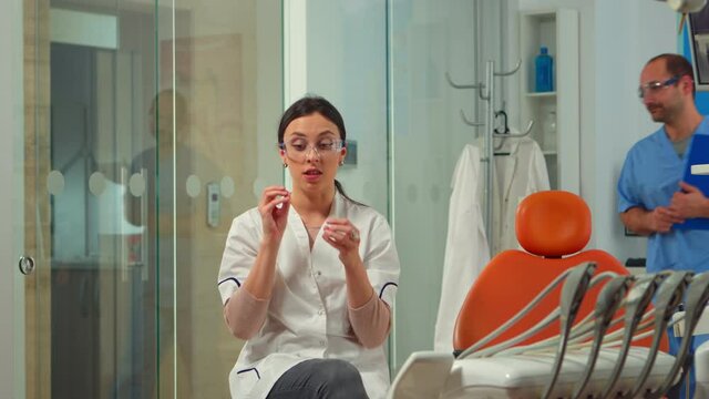 Woman stomatologist speaking on video camera sitting on chair in dental clinic with assistent in background. Dentistry doctor looking on webcam explaining treatment while nurse is talking with patient