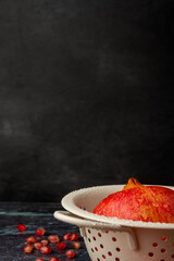 Close-up of white colander with wet red pomegranate, with seeds out of focus, on blue marble and black background, in vertical, with copy space