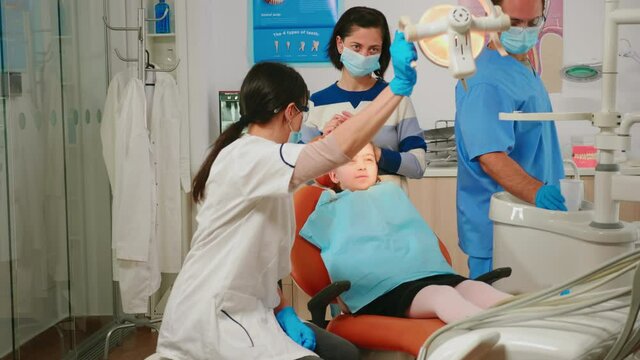 Woman dentist technician lighting the lamp for examining little patient. Orthodontist speaking to girl sitting on stomatological chair while nurse preparing tools for examination in modern clinic.