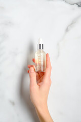 Transparent dropper bottle with serum lotion or essential oil in female hand over marble table. Skin care cosmetics concept.