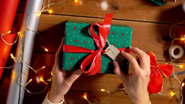 holidays, new year and christmas concept - woman holding gift box in green wrapping paper package with red bow and name tag at home over snow