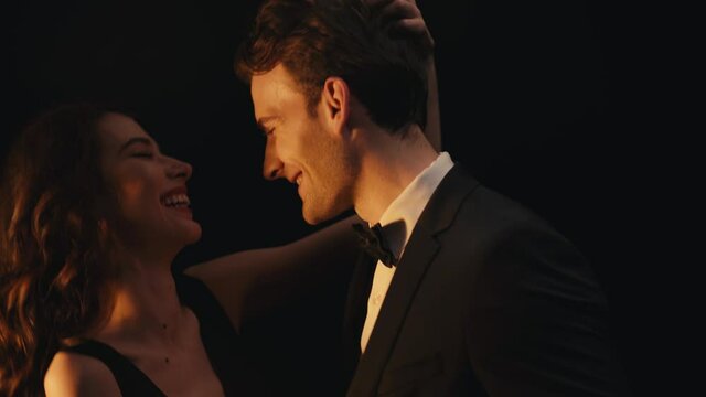 Happy elegant woman laughing while dancing with man in suit isolated on black