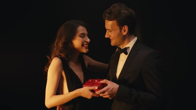 Surprised young woman receiving gift box from man in suit isolated on black