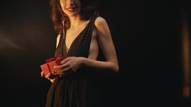 Cropped view of elegant smiling woman holding red gift box on black background