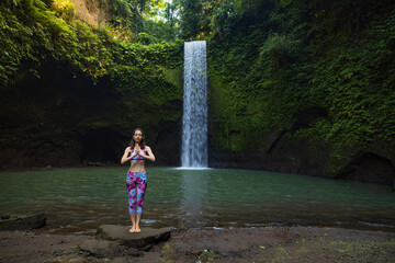 Woman standing on the stone, practicing yoga near waterfall. Hands in namaste mudra. Healthy life...