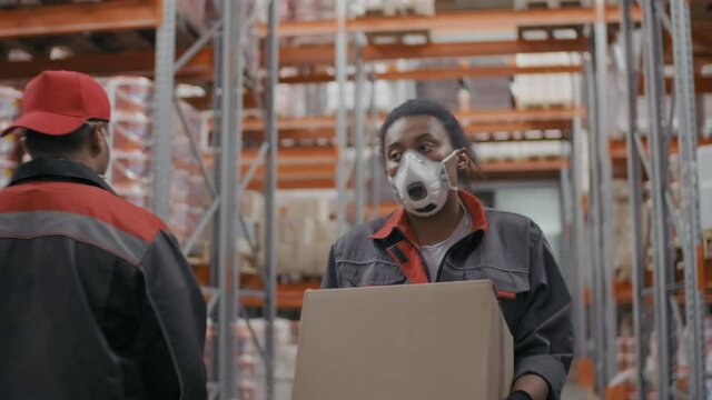 Medium slowmo footage of black woman in protective mask and construction uniform carrying big box walking along pallet racks at warehouse