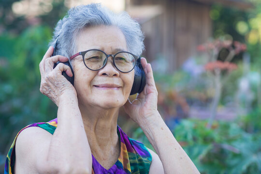 Portrait of a senior woman wearing wireless headphones listening to a favorite song while standing in a garden. Concept of aged people and relaxation