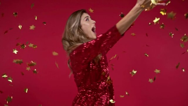 Slow motion of cheerful woman spinning near falling confetti on red background