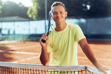 Young man teenager with tennis racket standing near net on clay court