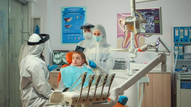 Pediatric dentist wearing protection suit treating girl patient in new normal stomatological unit showing teeth x-ray. Medical team wearing face shield coverall, mask, gloves, explaining radiography