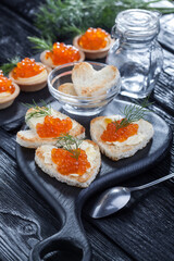 appetizer, heart-shaped snacks with butter and red caviar served with dill on a black wooden table