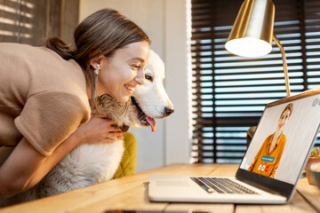 Young woman with a cute white dog working on a laptop, having a video call with colleque in the...