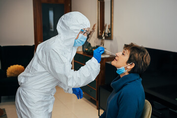 Health Professional in PPE, face mask and shield, introducing a nasal swab to a senior female patient on a home visit. Rapid Antigen Test to analyze nasal sampling for Covid-19, Coronavirus Pandemic. - obrazy, fototapety, plakaty