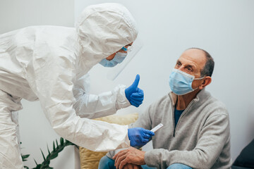 Health professional in a PPE suit, mask, face shield showing the patient the negative result for Covid-19. Antigen test to an elderly patient during a home visit. Coronavirus Pandemic.