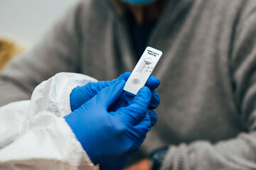 Close up of doctor hands with protective gloves and PPE suit, showing a test device to senior patient. Review of a Rapid Antigen Test for SARS Covid-19.