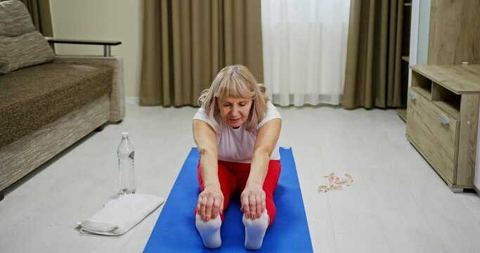 Elderly Woman Doing Stretching Exercises At Home Seated On A Mat In The Living Room Reaching Forwards To Touch Her Toes With A Smile For The Camera In A Health And Fitness Concept