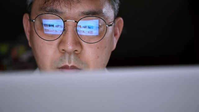An Asian middle-aged man eyes in eyeglasses late at night scrolling in front of the laptop. Eyeglasses with the reflection of the computer screen.