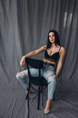 Photoshoot of a young brunette. The girl is sitting on a chair in jeans. Grey background.