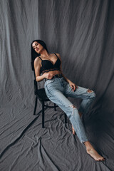 Obraz premium Photoshoot of a young brunette. The girl is sitting on a chair in jeans. Grey background.
