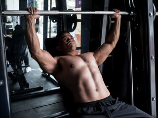 A muscular asian man performs incline bench presses on a Smith Machine at a gym or fitness center....