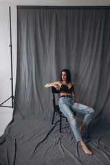 Photoshoot of a young brunette. The girl is sitting on a chair in jeans. Grey background.