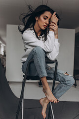 Photoshoot of a young brunette on a chair. Girl in jeans. Grey background.