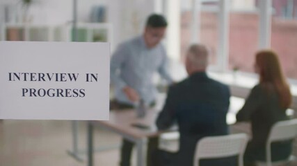 Interview in progress sign printed on paper hanging on glass wall in workspace, blurred people sitting in room on background