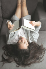 An Asian woman with messy hair lies down on a grey sofa with her feet up smiling and reading a book...