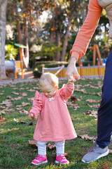 Adorable baby standing on grass, holding dad hand and looking down. Cropped unrecognizable father walking with little daughter in pink dress in autumn park. Fatherhood, holiday and weekend concept