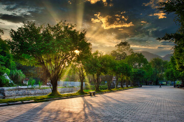 Sunset in the city of Hoa Lư in Vietnam creeping in through a long line of green trees while the light is reflected on the clouds above. 