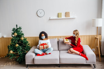 Celebrating Christmas and New Years 2021 during pandemic Covid-19 concept. Two multiethnic women in protective masks sitting on the couch and exchange gifts.