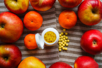 Red apples and tangerines lie around vitamins on a mottled light canvas, focus on scattered vitamins